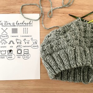May include: A gray knitted hat with a tag that says "This item is handmade! here's a little bit of information on it." The tag also includes icons for knitting, weaving, crocheting, different types of yarn, and washing instructions.