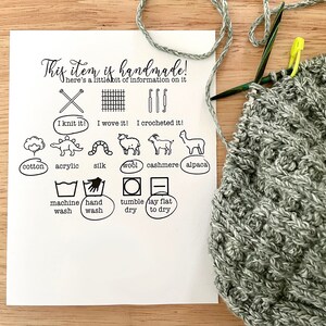 May include: A white card with black text and icons describing the handmade nature of the item. The card includes information about the materials used, such as cotton, acrylic, silk, wool, cashmere, and alpaca. It also includes care instructions, such as machine wash, hand wash, tumble dry, and lay flat to dry. The card is next to a gray knitted item.