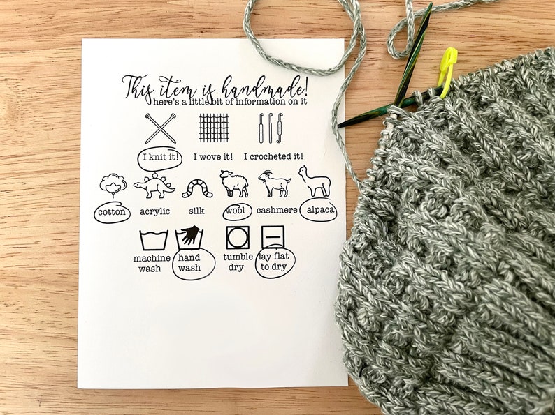 May include: A white card with black text and icons describing the handmade nature of the item. The card includes information about the materials used, such as cotton, acrylic, silk, wool, cashmere, and alpaca. It also includes care instructions, such as machine wash, hand wash, tumble dry, and lay flat to dry. The card is next to a gray knitted item.