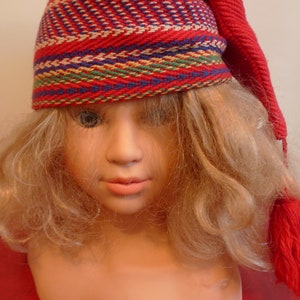 May include: A red knitted hat with a colourful striped pattern. The hat has a long tassel and is worn by a doll with blonde hair.