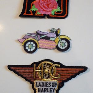 May include: Three embroidered patches featuring Harley Davidson motorcycle themes. The top patch is black with red trim and a pink rose. The middle patch is a pink and yellow sidecar motorcycle. The bottom patch is brown with gold trim and the text "Ladies of Harley".
