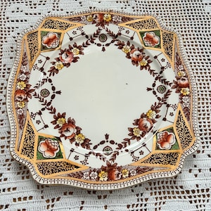 Può includere: Un piatto quadrato in ceramica bianca con un bordo ondulato. Il piatto presenta un motivo floreale marrone, oro e verde con un motivo geometrico attorno al bordo.