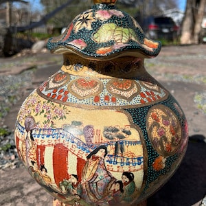 Può includere: Un grande vaso in ceramica decorato, con un coperchio dorato e un design floreale e geometrico colorato. Il vaso presenta una scena di persone in abiti tradizionali giapponesi.