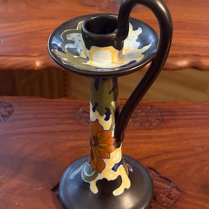 May include: A decorative ceramic candlestick holder with a dark brown base and handle. The candlestick features a floral design in orange, yellow, and blue against a green and brown background. The handle curves gracefully from the top.