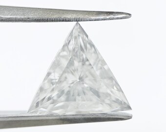 Puede incluir: Un diamante transparente de corte triangular sostenido por unas pinzas plateadas. La gema tiene una superficie facetada, que refleja la luz y muestra su forma geométrica. El diamante está sobre un fondo blanco brillante.