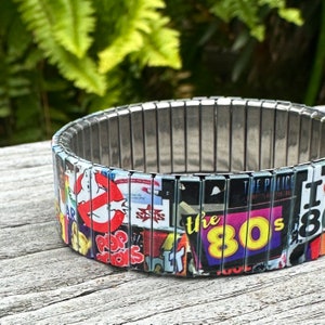 May include: A silver expandable bracelet with a colorful collage of 1980s pop culture icons, including Ghostbusters, The Police, and the text "the 80s".