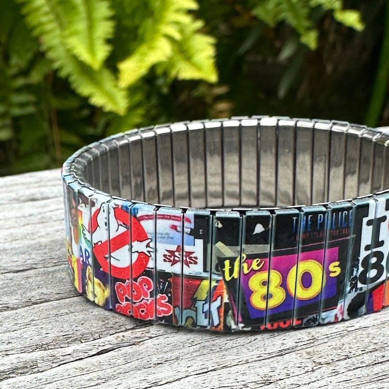 May include: A silver expandable bracelet with a colorful collage of 1980s pop culture icons, including Ghostbusters, The Police, and the text "the 80s".