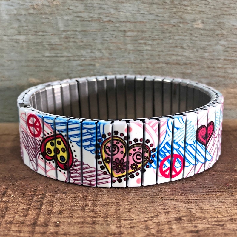 Op de afbeelding: Een witte uitbreidbare armband met een kleurrijk ontwerp met vredestekens, harten en vleugels. De armband is gemaakt van metaal en heeft een zilveren afwerking.
