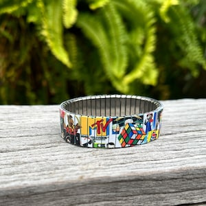 May include: A colorful bracelet with a silver metal band and a design featuring images from the 1980s, including MTV, a Rubik's Cube, and a cartoon character.