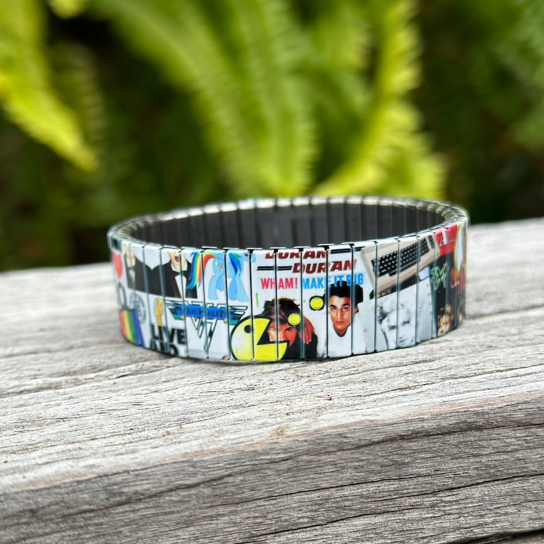 May include: A silver expandable bracelet with a collage of 80s music album covers. The covers feature bands like Duran Duran, Wham!, and Def Leppard. The bracelet is made of metal links and has a flexible design.