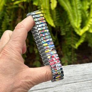 May include: A silver watch band with a colorful, patterned design. The band is made up of individual links, each with a different image. The images are mostly abstract and geometric, with some featuring cartoon characters.