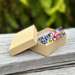 May include: A colorful bracelet made of small squares with images from the 80s, including a Rubik's Cube, Ghostbusters, and the Coca-Cola logo. The bracelet is in a brown cardboard box.