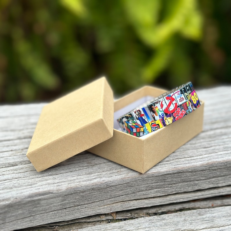 May include: A colorful bracelet made of small squares with images from the 80s, including a Rubik's Cube, Ghostbusters, and the Coca-Cola logo. The bracelet is in a brown cardboard box.