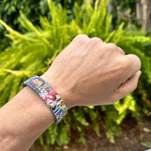May include: A colorful bracelet made of multiple small squares with different patterns and colors. The bracelet is made of metal and has a flexible design.
