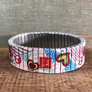 Op de afbeelding: Een kleurrijke armband met een witte achtergrond en verschillende ontwerpen, waaronder harten, vleugels en vredestekens. De armband is gemaakt van metaal en heeft een magnetische sluiting. De tekst "Wrist Art" is op de armband gedrukt.