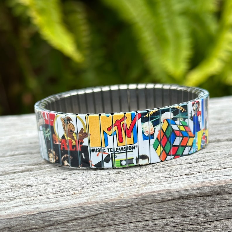 May include: A colorful bracelet with a silver metal band and images of popular 80s and 90s icons, including MTV, Alf, and a Rubik's Cube.