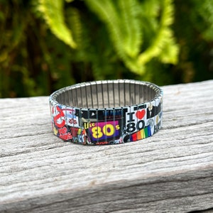 May include: A silver expandable bracelet with a colorful design featuring 80s pop culture icons, including the Ghostbusters logo, "The 80s" text, and "I Love the 80s" text.