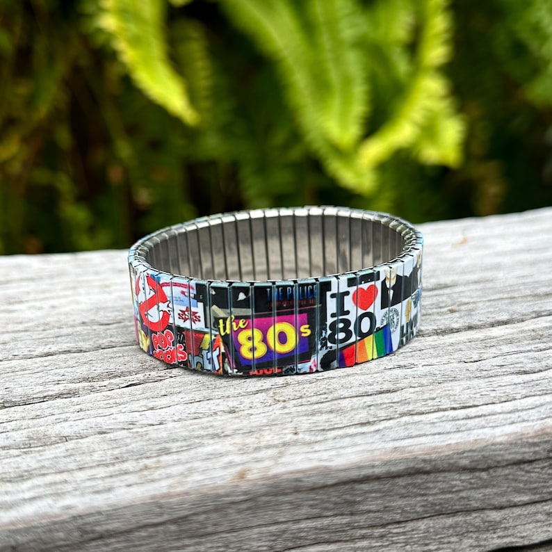 May include: A silver expandable bracelet with a colorful design featuring 80s pop culture icons, including the Ghostbusters logo, "The 80s" text, and "I Love the 80s" text.