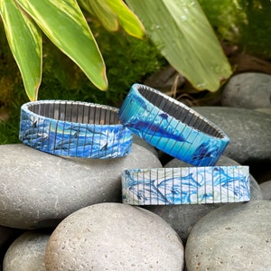 May include: Three blue and white dolphin patterned expandable bracelets. The bracelets are made of metal and have a stretchy design. The bracelets are on a bed of gray rocks.