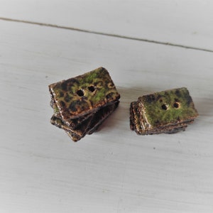 May include: A collection of rectangular ceramic buttons with two holes each. The buttons have a mottled green and brown glaze, stacked in small piles. The background is a white, textured surface.