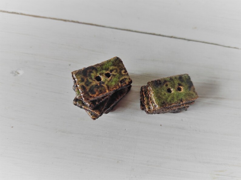 May include: A collection of rectangular ceramic buttons with two holes each. The buttons have a mottled green and brown glaze, stacked in small piles. The background is a white, textured surface.