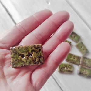 Può includere: Bottoni rettangolari in ceramica con una superficie strutturata verde muschio e marrone. Ogni bottone ha due fori per cucire. Sono visibili diversi bottoni, che suggeriscono un set per l'artigianato o l'abbellimento di capi.