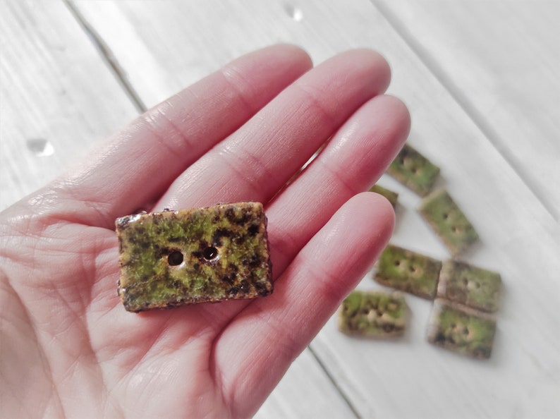 May include: Rectangular ceramic buttons with a textured, mossy green and brown surface. Each button has two holes for sewing. Several buttons are visible, suggesting a set for crafting or garment embellishment.