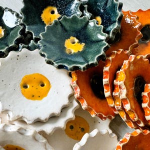 Puede incluir: Una colección de botones de cerámica en varios colores y diseños. Los botones son redondos y tienen un centro elevado con un pequeño agujero en el medio. Algunos botones son blancos con centros amarillos, otros son naranjas con bordes rojos y algunos son verdes con acentos azules.