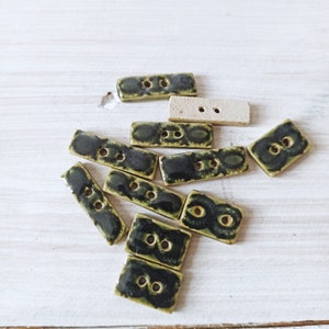 Puede incluir: Una colección de botones de cerámica verde y negro con un diseño floral. Los botones son rectangulares y tienen dos agujeros.