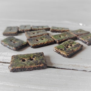 May include: A collection of rectangular ceramic buttons with two holes each. The buttons have a mottled green and brown glaze, giving them a rustic appearance. The buttons are scattered on a light gray surface.