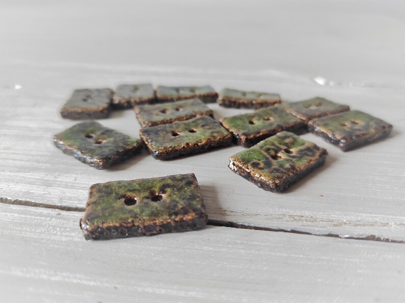 May include: A collection of rectangular ceramic buttons with two holes each. The buttons have a mottled green and brown glaze, giving them a rustic appearance. The buttons are scattered on a light gray surface.