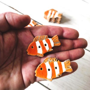 May include: Two orange and white ceramic fish buttons with black eyes. The fish have white stripes and are about 2 inches long.