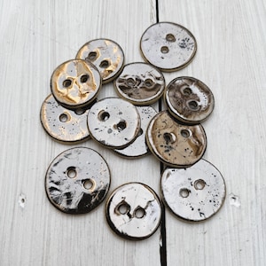 Puede incluir: Una colección de 11 botones redondos de cerámica en tonos marrones y dorados con un acabado metálico. Los botones tienen dos agujeros cada uno y están dispuestos en un patrón aleatorio.