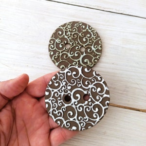 May include: Two large, round brown buttons with an embossed, swirling white design. Each button has two holes for sewing. The buttons are held in a hand, showcasing the size and detail of the decorative elements.