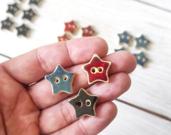 May include: Star-shaped buttons in blue, red, and black with gold trim. The buttons have two holes for sewing. Several other star buttons are scattered in the background on a white wooden surface.