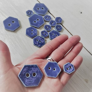 Può includere: Una collezione di bottoni in ceramica blu di diverse dimensioni, con forma esagonale e due fori. I bottoni hanno una superficie testurizzata e macchiata.