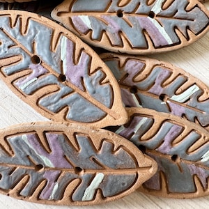 Può includere: Un set di sei bottoni in ceramica a forma di foglia con un design dipinto in grigio e viola. I bottoni hanno un foro al centro per cucire.
