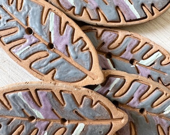 Könnte beinhalten: Sechs Keramik-Knöpfe in Blattform mit einem grau-violetten Design. Die Knöpfe haben ein Loch in der Mitte zum Nähen.