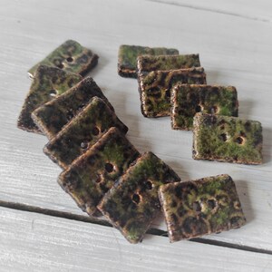 May include: A collection of square ceramic buttons with a mottled green and brown glaze. Each button has two holes for sewing. The buttons are arranged on a light-colored wooden surface.