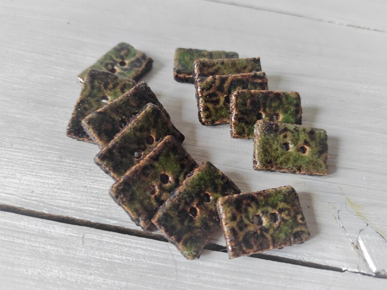 May include: A collection of square ceramic buttons with a mottled green and brown glaze. Each button has two holes for sewing. The buttons are arranged on a light-colored wooden surface.