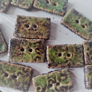 May include: A collection of rectangular ceramic buttons with two holes each. The buttons have a mottled green and brown glaze, giving them a rustic appearance. The edges are slightly rough, adding to the handmade aesthetic. Ideal for sewing and craft projects.