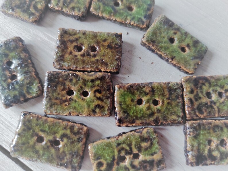 May include: A collection of rectangular ceramic buttons with two holes each. The buttons have a mottled green and brown glaze, giving them a rustic appearance. The edges are slightly rough, adding to the handmade aesthetic. Ideal for sewing and craft projects.