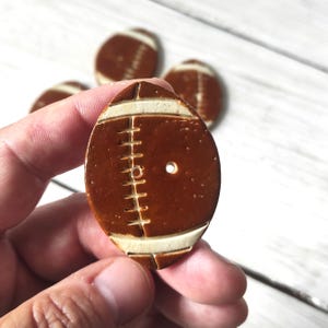 Puede incluir: Botones de cerámica con forma de balón de fútbol, marrón y crema. Los botones tienen rayas blancas y detalles de costura, con un agujero para coser. Se ven varios botones, uno de ellos en una mano.