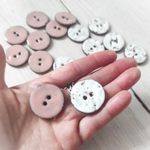 Könnte beinhalten: Eine Sammlung von Keramikknöpfen in Rosa und Weiß mit gesprenkeltem Glasur. Einige Knöpfe werden in einer Hand gehalten.