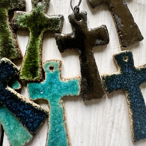 May include: A collection of ceramic cross ornaments in various colors, including green, blue, and black. The crosses are all different sizes and shapes, and some have a textured finish.