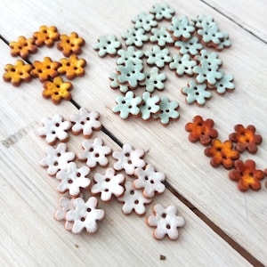 Könnte beinhalten: Sortiment an Keramik-Knöpfen in Blumenform in verschiedenen Farben, darunter Weiß, Hellblau und Orange. Die Knöpfe sind in drei Gruppen auf einer hellbraunen Holzoberfläche angeordnet.