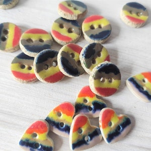 Può includere: Una collezione di bottoni in ceramica colorati in varie forme, tra cui forme rotonde e a forma di cuore. I bottoni presentano un design a strisce con colori giallo, rosso e blu.