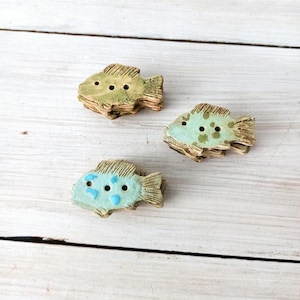 Può includere: Tre bottoni in ceramica a forma di pesce nei toni del verde e del blu. Ogni bottone ha tre fori e un design strutturato e stratificato. I pesci sono disposti su una superficie di legno bianco.
