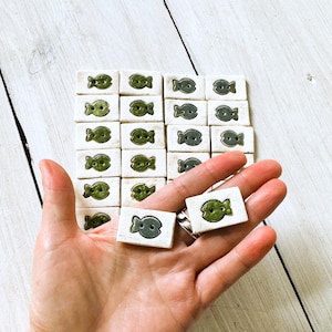 Puede incluir: Una colección de 24 botones de cerámica con forma de pez. Los botones son blancos con un diseño de pez verde. Dos botones se muestran en la mano, y el resto en una cuadrícula.