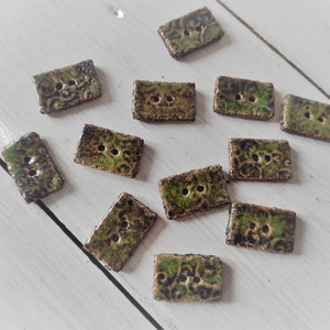 May include: A collection of rectangular ceramic buttons with two holes each. The buttons have a mottled green and brown glaze with a textured, patterned surface. They are scattered on a white wooden surface.